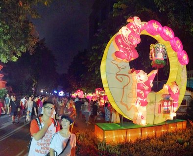 Night scene of a large illuminated crescent moon lantern display with traditional Chinese figures and pink lanterns at a Mid-Autumn Festival celebration with crowds of people walking on a tree-lined street in Guangzhou, China