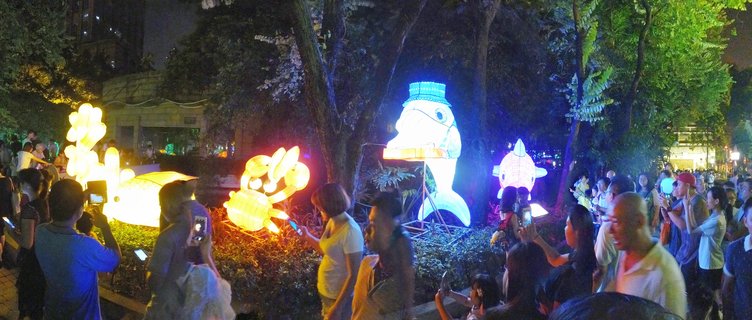 Night scene showing multiple illuminated fish-shaped lanterns in yellow, white, and orange colors with a crowd of people photographing the display in an urban park in Guangzhou, China