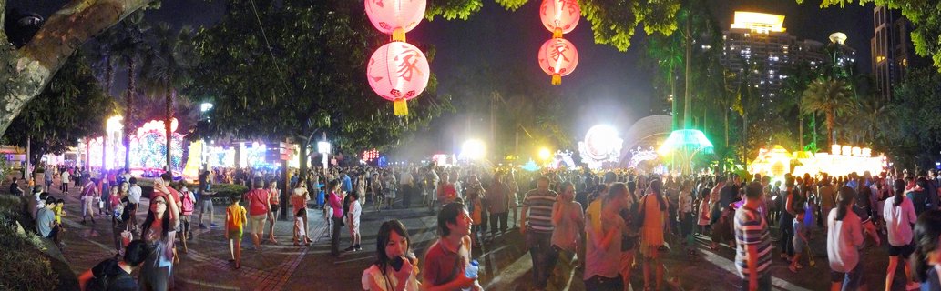 Night scene of a crowded street festival in Guangzhou with red Chinese lanterns hanging from trees, bright illuminated displays, and many people walking on a paved pedestrian area with modern buildings in the background