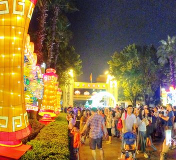 Night scene showing large illuminated yellow and red Chinese lanterns with geometric patterns, crowds of people walking on a pathway, palm trees, and a decorated entrance gate with Chinese characters and hanging red lanterns