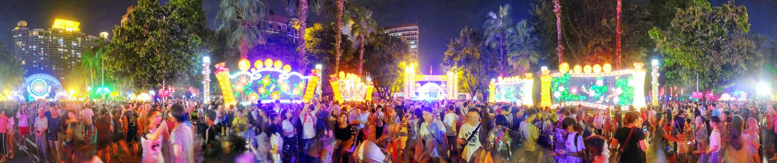 Night panoramic photograph of a crowded outdoor lantern festival with colorful illuminated displays in purple, yellow, and green, palm trees, and city buildings in the background