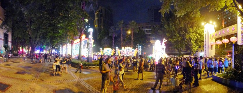 Night photograph of a crowded public plaza in Guangzhou during a Chinese lantern festival, with illuminated traditional lantern displays, red hanging lanterns at an entrance gate, trees, and visitors walking on brick pavement