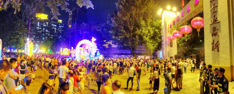 Night festival scene showing large crowd of people gathered on pedestrian plaza with traditional red Chinese lanterns hanging from building, illuminated parade float with purple and blue lights, trees, and modern buildings in background