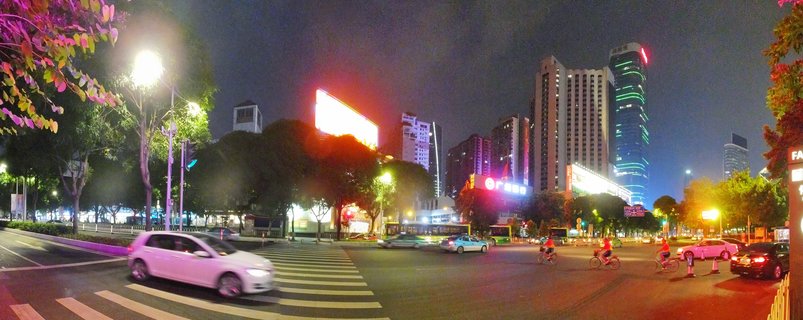 Night panoramic cityscape of Guangzhou showing illuminated skyscrapers, street intersection with moving vehicles, cyclists, crosswalk, trees, and colorful neon signs against dark sky