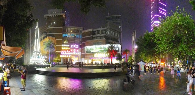 Night photograph of a busy outdoor shopping plaza in Guangzhou with an illuminated Eiffel Tower replica, colorful building signage, pedestrians walking on wet pavement, and a tall skyscraper with purple LED lighting