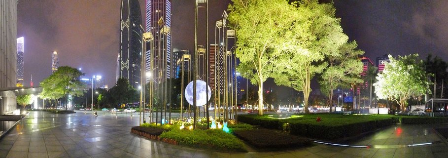 Night photograph of Guangzhou city plaza with illuminated spherical moon art installation, modern skyscrapers including Guangzhou IFC tower, uplit trees, and wet reflective pavement