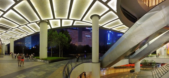 Night view of a modern shopping plaza with illuminated geometric ceiling panels, white columns, curved escalators, and pedestrians walking with illuminated skyscrapers in the background