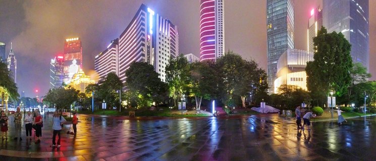 Nighttime panoramic view of illuminated skyscrapers in Guangzhou China with colorful building lights reflecting on wet pavement while people walk across a public plaza