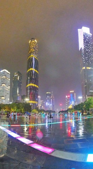 Night cityscape showing Guangzhou IFC tower with golden light bands and surrounding skyscrapers reflecting on wet pavement with colorful LED ground lights and people walking with umbrellas