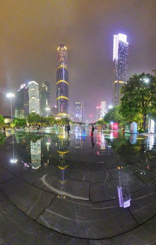 Night photograph of illuminated skyscrapers in Guangzhou's financial district with building lights reflecting on wet stone plaza where people are walking