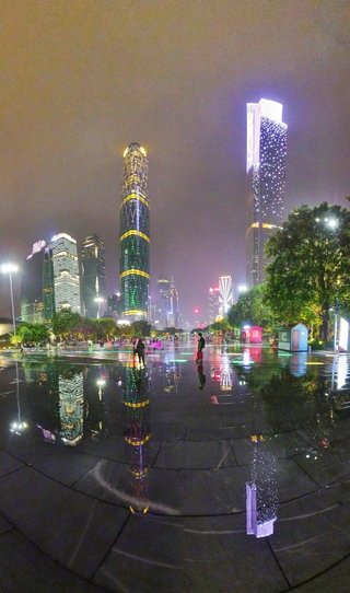 Night cityscape showing illuminated skyscrapers including Guangzhou IFC and CTF Finance Centre with colorful reflections on wet pavement and people walking in foreground plaza