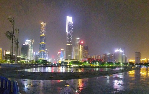 Night photograph of Guangzhou city skyline with illuminated skyscrapers including cylindrical and rectangular towers, reflections visible on wet ground in foreground