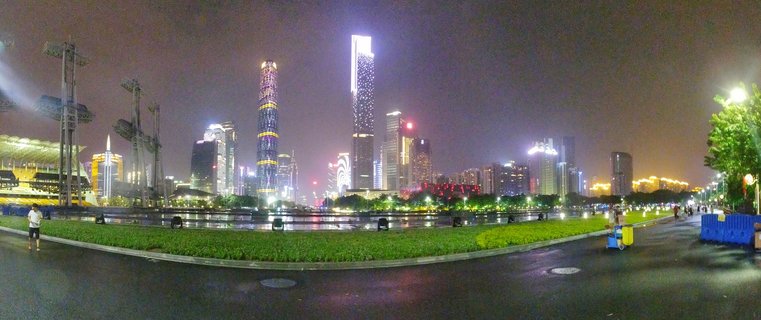 Night panoramic view of Guangzhou's illuminated skyscraper skyline including the CTF Finance Centre, viewed from a waterfront promenade with green landscaping and wet pavement reflecting city lights