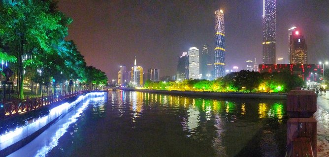 Night photograph of Guangzhou city skyline with illuminated skyscrapers including CTF Finance Centre reflecting in Pearl River water, trees with green uplighting along waterfront promenade