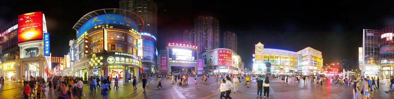 Panoramic night photograph of a crowded pedestrian shopping plaza in Guangzhou with illuminated multi-story commercial buildings, neon signs, and people walking on paved walkways