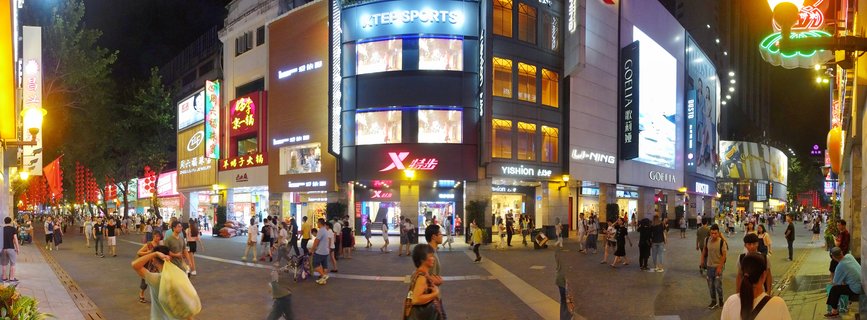 Wide night photograph of a crowded pedestrian shopping street in Guangzhou with illuminated storefronts, neon signs, and people walking on paved plaza