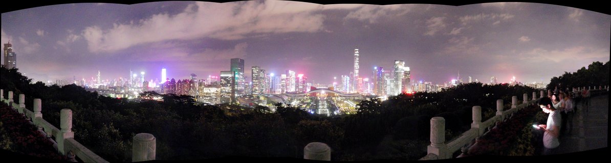 Panoramic night photograph of Shenzhen city skyline with illuminated skyscrapers, cloudy purple-tinted sky, dark trees in foreground, and stone railing with visitors on viewing platform