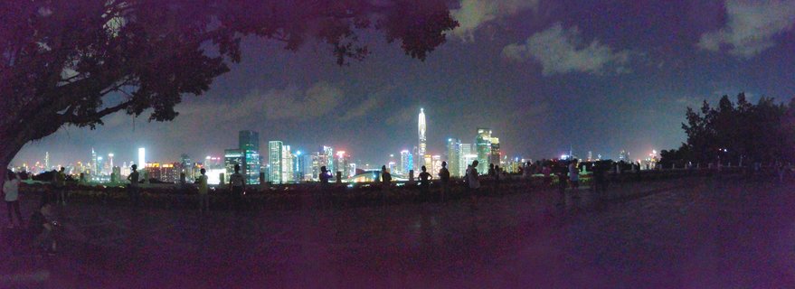 Panoramic night photograph of Shenzhen city skyline with illuminated skyscrapers, silhouettes of people on waterfront promenade, trees framing left and right sides