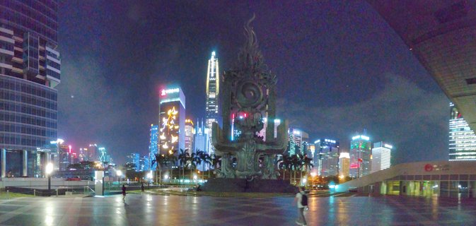 Night photograph of Shenzhen city center showing illuminated skyscrapers behind a large ornate bronze sculpture in a paved public plaza with one pedestrian walking