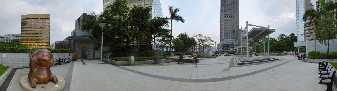 Bronze pig sculpture on a plaza in Hong Kong with tall buildings, palm trees, benches, and people walking in the background under cloudy sky