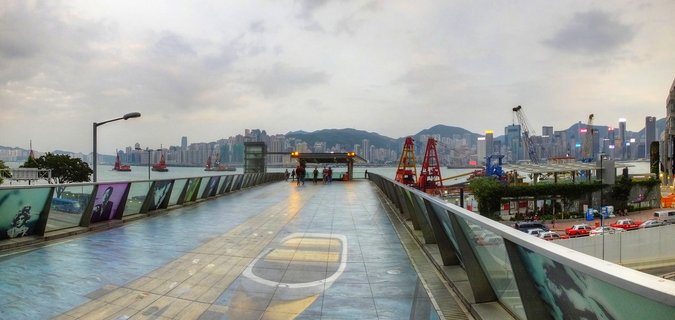 Wide waterfront promenade in Hong Kong with glass railings, people walking, Victoria Harbour with red junk boats, and city skyline with mountains in background under cloudy sky