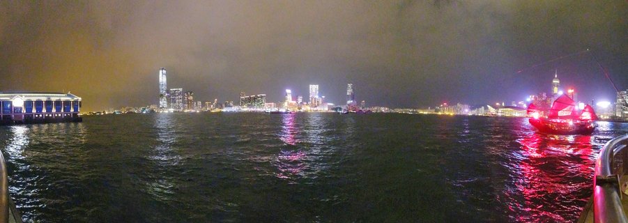 Nighttime panoramic view of Hong Kong Victoria Harbour skyline with illuminated skyscrapers, traditional red junk boat, and city light reflections on water