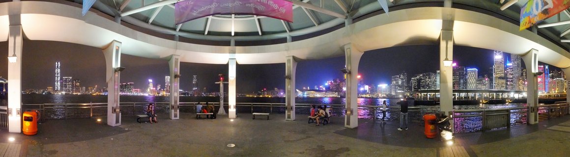 Night view of Hong Kong's illuminated skyline across Victoria Harbour from a covered waterfront promenade with white pillars, where people sit on benches watching the city lights reflect on the water