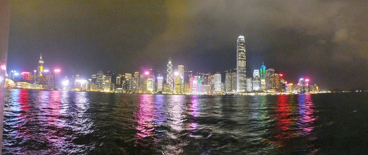 Panoramic night photograph of Hong Kong's Victoria Harbour skyline showing illuminated skyscrapers with colorful light reflections on water