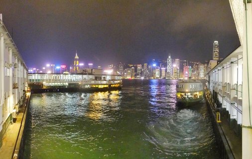 Night photograph of Hong Kong's Victoria Harbour showing illuminated skyscrapers on Hong Kong Island, two Star Ferry boats at piers, and colorful light reflections on water
