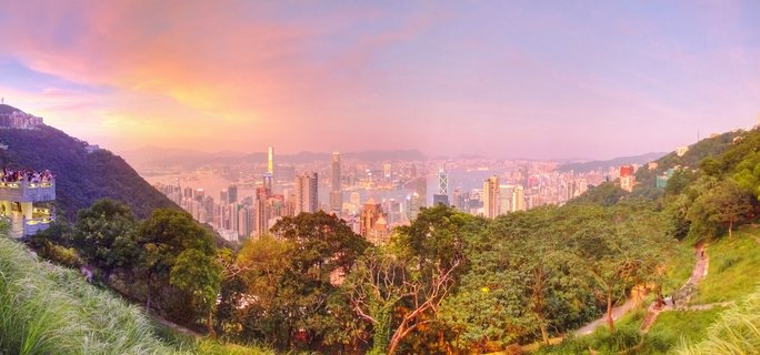 Panoramic cityscape of Hong Kong at sunset viewed from Victoria Peak, showing dense skyscraper skyline across Victoria Harbour with green mountain slopes and walking paths in the foreground under pink and orange sky