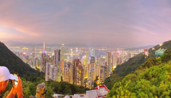 Panoramic view of Hong Kong's illuminated skyscraper skyline at twilight from Victoria Peak, with Victoria Harbour visible between densely packed buildings and green hillsides framing the scene