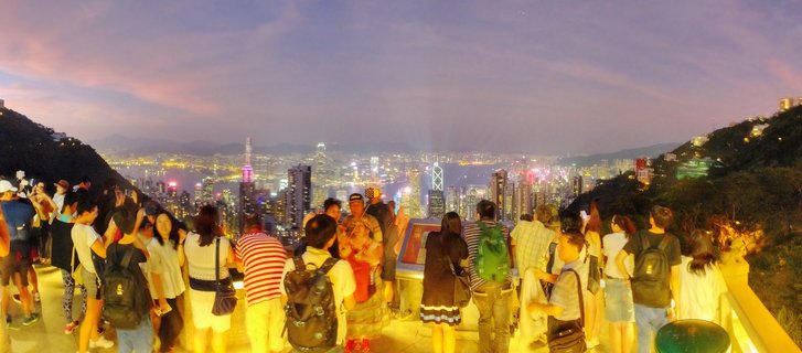 Panoramic view of tourists on an observation deck at Victoria Peak overlooking Hong Kong's illuminated skyline and Victoria Harbour at dusk with purple and pink sky