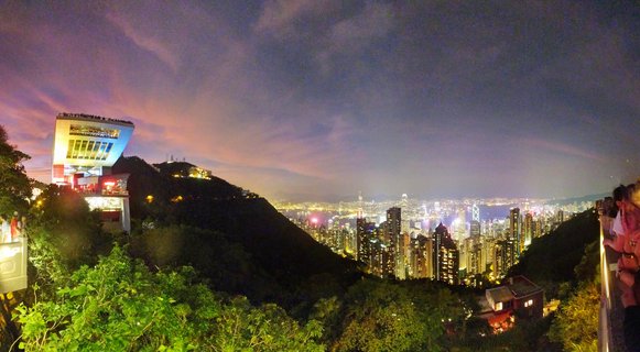 Nighttime panoramic view of Hong Kong's illuminated skyline from Victoria Peak showing skyscrapers, Victoria Harbour, Peak Tower building, and tourists at viewing platforms under a purple twilight sky