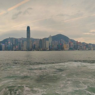 Hong Kong city skyline with tall skyscrapers including IFC Tower viewed across Victoria Harbour water with boat wake in foreground and Victoria Peak mountain in background under cloudy sky