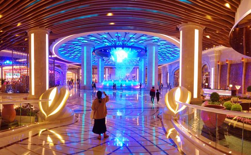 Interior view of a luxury casino resort lobby in Macau featuring a central blue-lit fountain, marble columns with vertical lighting, polished reflective floors, and visitors walking through the space