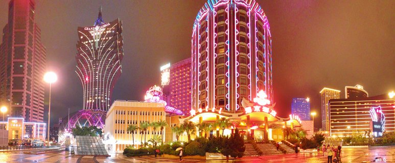 Night cityscape of Macau showing the Grand Lisboa casino tower with distinctive lotus-shaped design and Hotel Lisboa with pink neon vertical lighting, wet plaza with pedestrians and palm trees in foreground