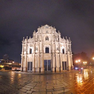 The illuminated stone facade of the Ruins of St. Paul's in Macau at night, featuring baroque architectural details and religious statues, with wet cobblestone ground reflecting warm street lamp light and several tourists visible on the right side of the plaza