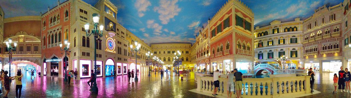 Wide interior panorama of The Venetian Macao showing colorful Italian-style building facades, painted sky ceiling, ornate street lamps, shoppers walking on reflective floors, and a bridge over an indoor canal
