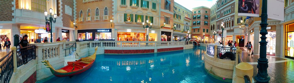 Indoor canal at Venetian Macao with red gondola, Italian-style buildings, shoppers on walkways, and decorative street lamps