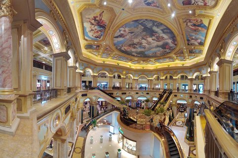Multi-level luxury shopping mall interior with ornate baroque ceiling frescoes, gold columns, marble floors, and escalators in Macau casino resort