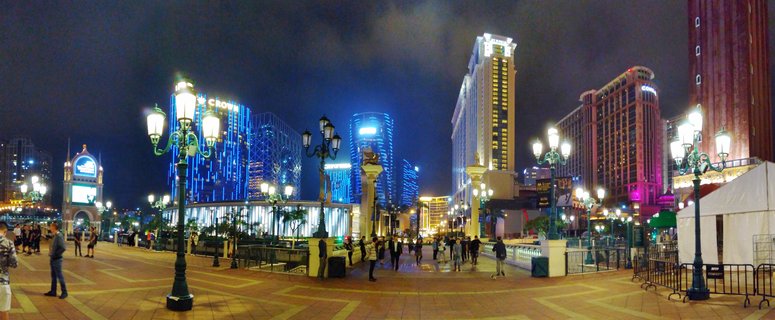 Night panoramic view of Macau's Cotai Strip showing illuminated casino resorts including Crown Towers with blue LED lighting, ornate street lamps, and pedestrians on a paved plaza