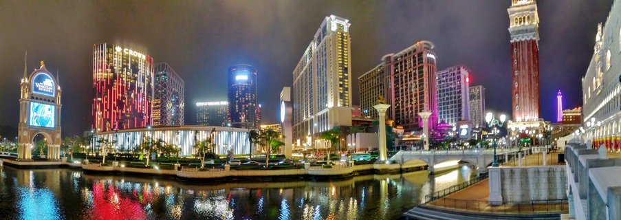 Night panoramic view of Macau Cotai Strip showing illuminated casino resort buildings with colorful lights reflecting in water canal