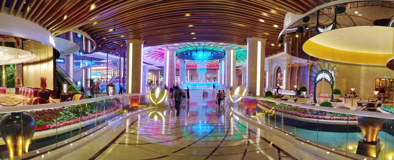 Interior view of a luxury casino resort atrium with colorful LED ceiling lights, marble floors, columns, glass railings, and people walking through the space