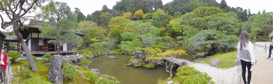 Panoramic view of Ginkaku-ji Temple's zen rock garden with raked white sand patterns in curved lines, traditional Japanese wooden buildings with dark tiled roofs, pine trees, and visitors walking on pathways under overcast sky