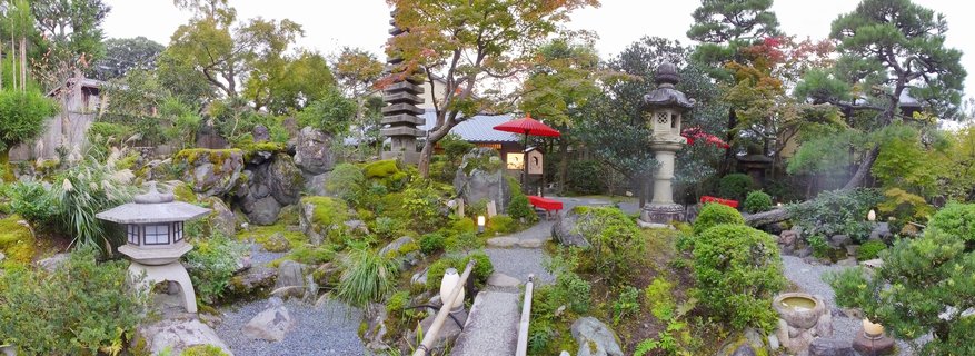 Panoramic view of a traditional Japanese inn with wooden architecture and large glass doors showing people inside, next to a Japanese garden with stone lanterns, manicured trees, rocks, and a stone pathway