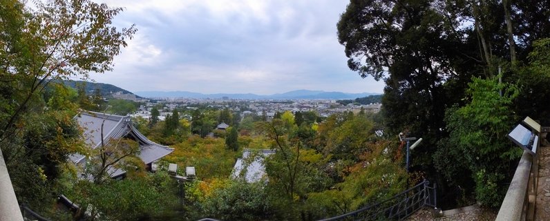 Panoramic view of Kyoto from temple showing traditional Japanese roofs, autumn trees, city buildings, and mountains under cloudy sky