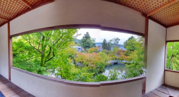 Interior view from a traditional Japanese building with wooden lattice ceiling looking out through a large curved window at a garden with pond, bridge, and trees