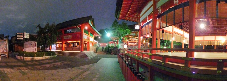 Traditional Japanese shrine buildings with orange-red vermilion walls and curved dark roofs illuminated at night, stone pathway leading through temple complex in Kyoto Japan