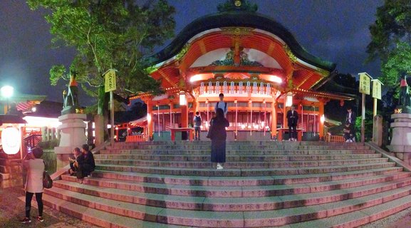 Night photograph of an illuminated traditional Japanese shrine building with curved roof, orange-red painted wood, stone staircase with visitors, fox statues on pedestals, and trees against dark blue evening sky