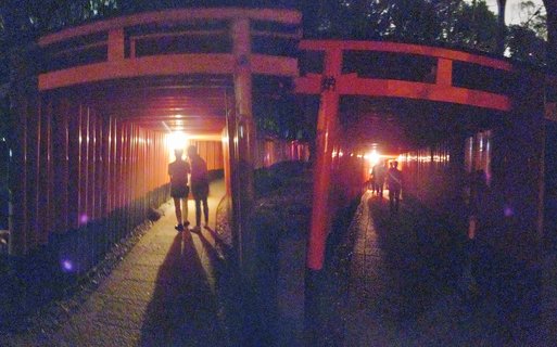 Night photograph showing people walking through illuminated orange-red torii gate tunnels at Fushimi Inari Shrine in Kyoto, Japan, with long shadows cast on the pathway